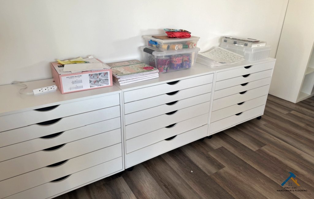 Custom white storage cabinets installed in an Eagle Mountain, Utah craft room with newly updated vinyl plank flooring; organized workspace showing professional cabinetry assembly and handyman services by Fixi-LLC.